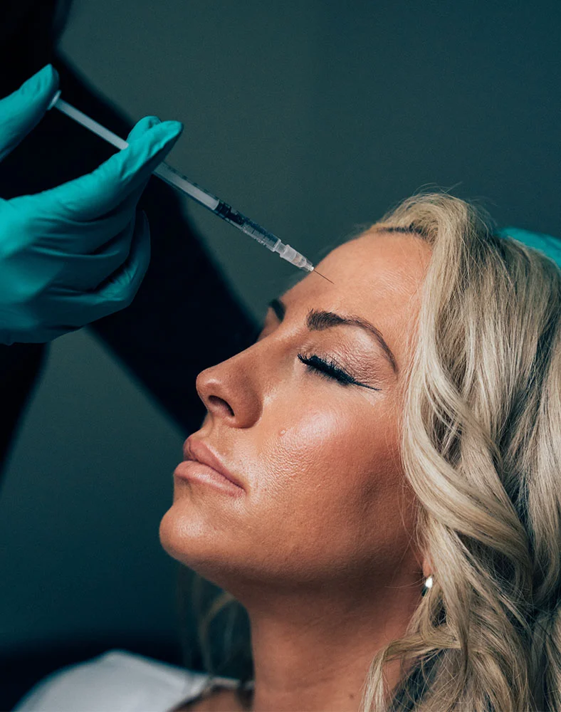Close-up of a woman with eyes closed receiving a cosmetic injection to her forehead. A person wearing teal medical gloves holds a clear syringe near the injection site. The dark background emphasizes the woman's face and the injection area, creating a focused, clinical atmosphere. - Botox and Wrinkle Relaxers in Carmel, IN