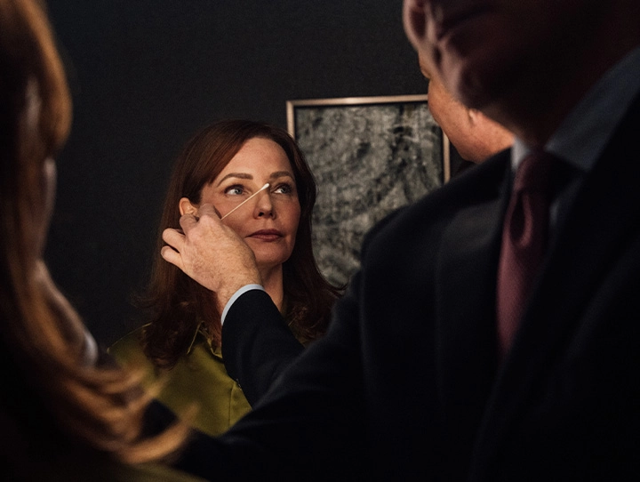Mid-shot of a consultation. Dr. Hamilton, in a dark suit and maroon tie, examines a woman’s face using a thin, light-colored tool near her nose and brow. The woman, with reddish-brown hair and wearing an olive green top, looks slightly upward. Focused lighting highlights their faces and hands against a dark background with framed art. - Brow Lift in Carmel, IN