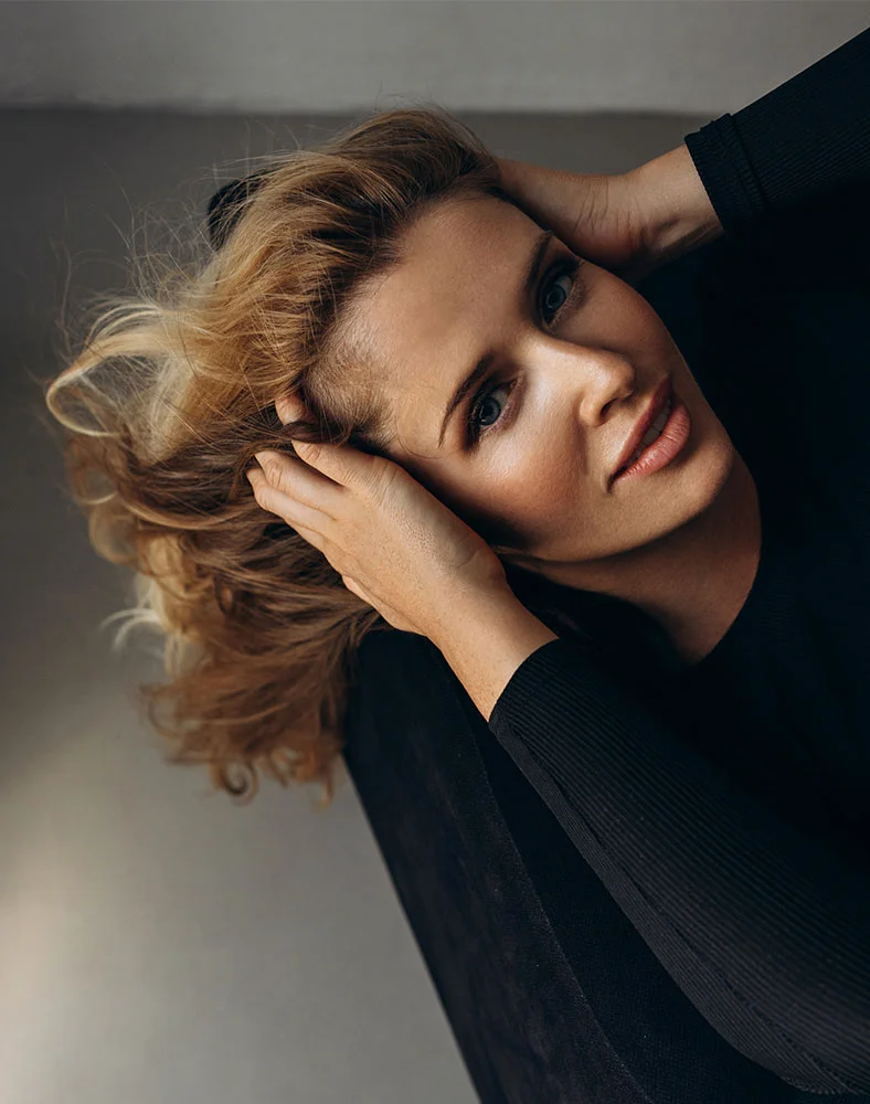 High-angle close-up of a light-skinned woman with slightly tousled, medium-length light brown or blonde hair. She gently holds her head with both hands, fingers resting on either side in a relaxed, contemplative pose. Her expression is serene as she looks directly at the camera. She has light blue eyes and natural pink lips, and wears a close-fitting, ribbed black long-sleeve top. The background is a neutral light gray or beige wall, softly lit with diffused lighting that highlights her features and conveys a calm, feminine atmosphere. - Browlift in Carmel, IN