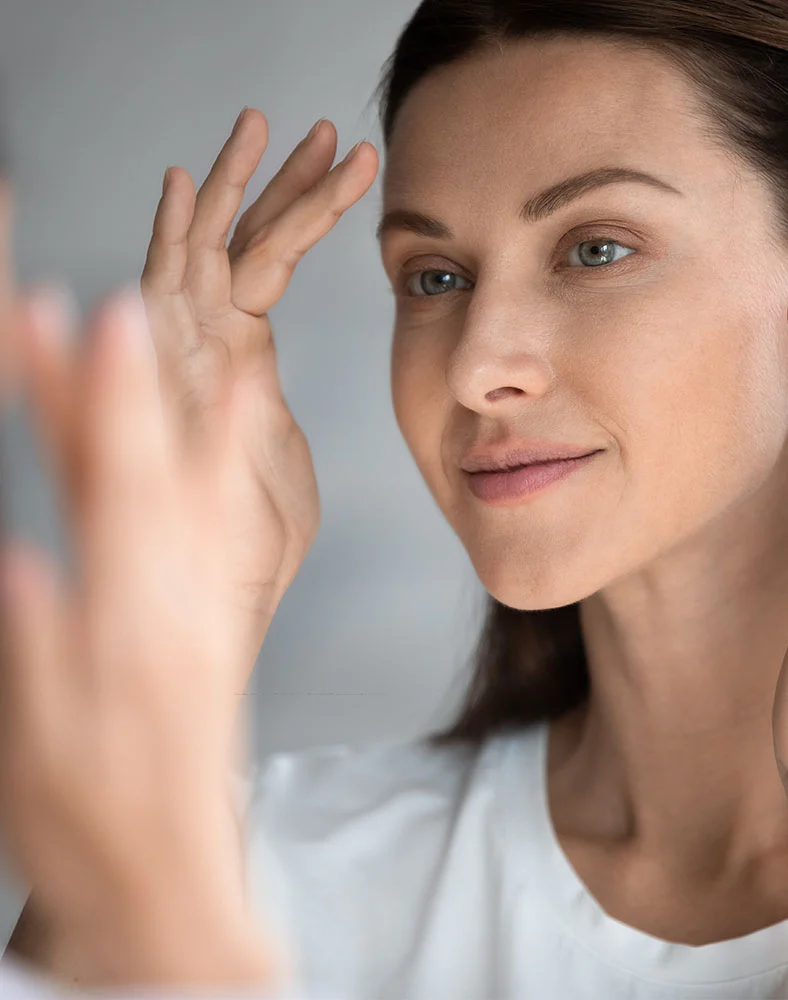 A woman with dark hair, wearing a white shirt, gently touches her forehead and looks thoughtfully off into the distance. The lighting is soft and the backgroud is a neutral, light gray. - Diamond Glow in Carmel, IN