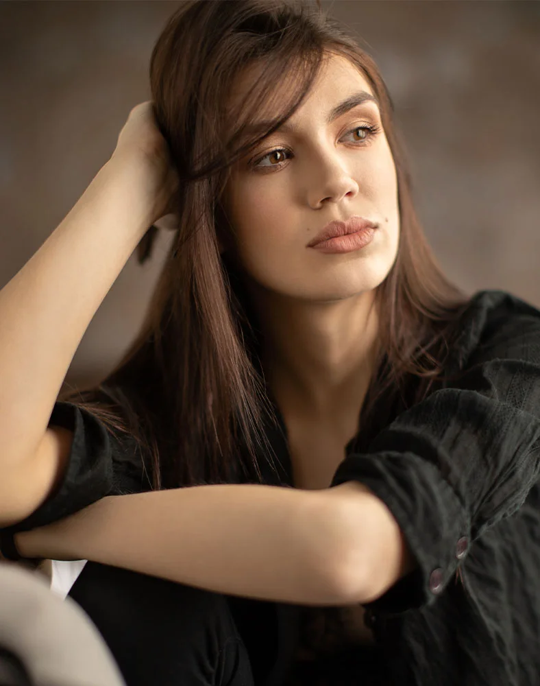 A portrait shot of a woman with long brown hair, gazing thoughtfully off to the side. She's wearing a dark-colored collared shirt and a serene expression. The lighting is soft and flattering. - Facial Fat Grafting including Nanofat in Carmel, IN