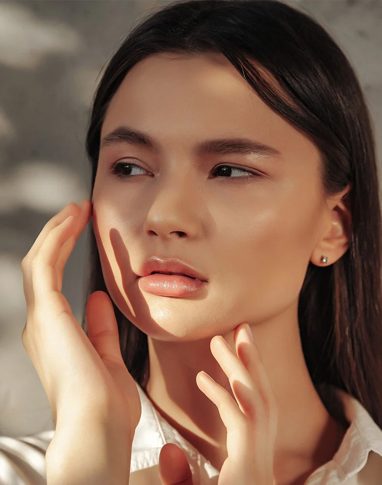 Close-up of a young woman with a light complexion and long dark brown hair, gazing softly to the side with a thoughtful expression. Natural sunlight highlights her smooth, glowing skin, while subtle shadows define her features. Her hands rest gently on her face and neck, and she wears small stud earrings. A white shirt is visible, and the blurred background features dappled sunlight on a light gray surface. - Hydrafacial in Carmel, IN