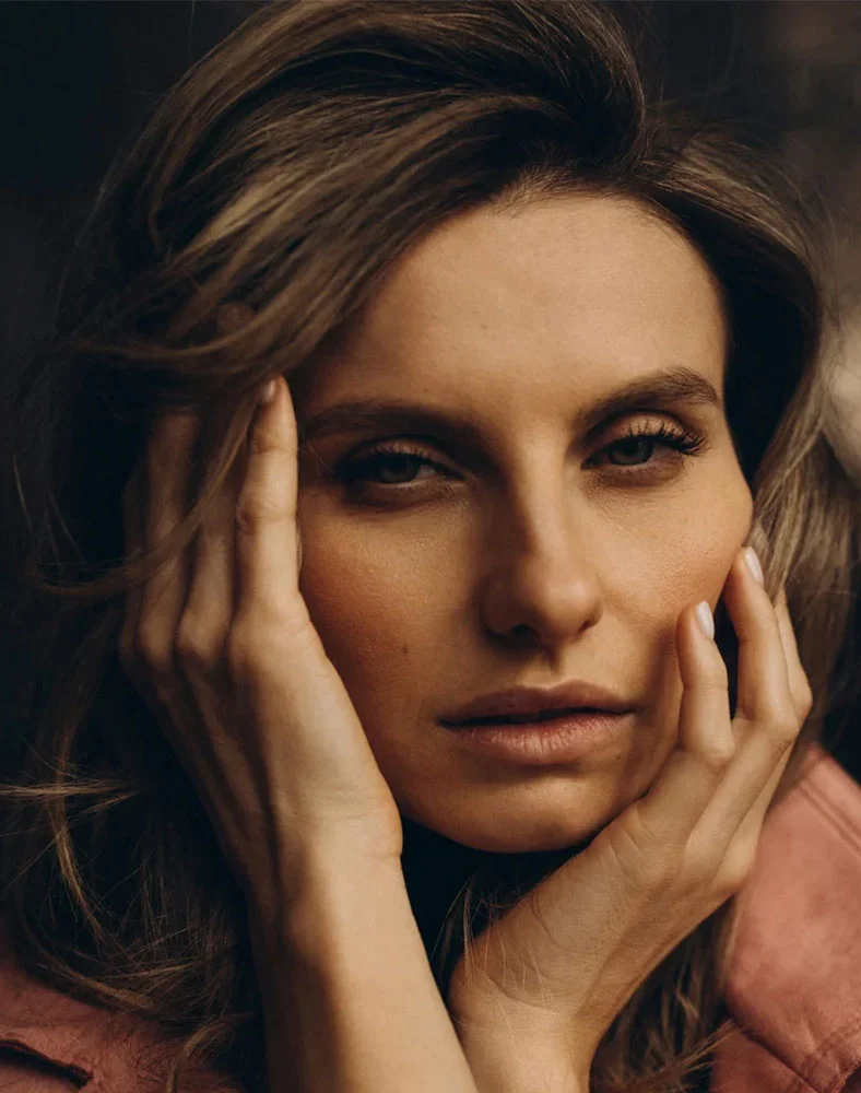 Close-up of a woman with shoulder-length, wavy light brown hair, gazing downward with a contemplative expression. Her light-toned skin is smooth, and she wears subtle makeup with soft eyeshadow and light lipstick. Her hands gently rest on her temples, and a dusky rose-colored top is visible. Soft lighting highlights her facial features against a neutral background, adding a touch of drama. - Laser Skin Resurfacing in Carmel, IN