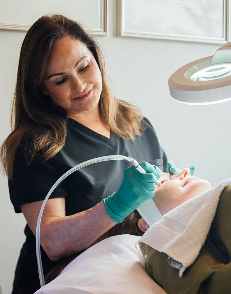 A woman in a dark scrub top performs a facial treatment on a reclining client covered with a white towel. She wears teal gloves and uses a small, light device with a tube on the client’s face. A round magnifying light hovers above. The background features a light wall with framed art. - Microneedling Skinpen in Carmel, IN
