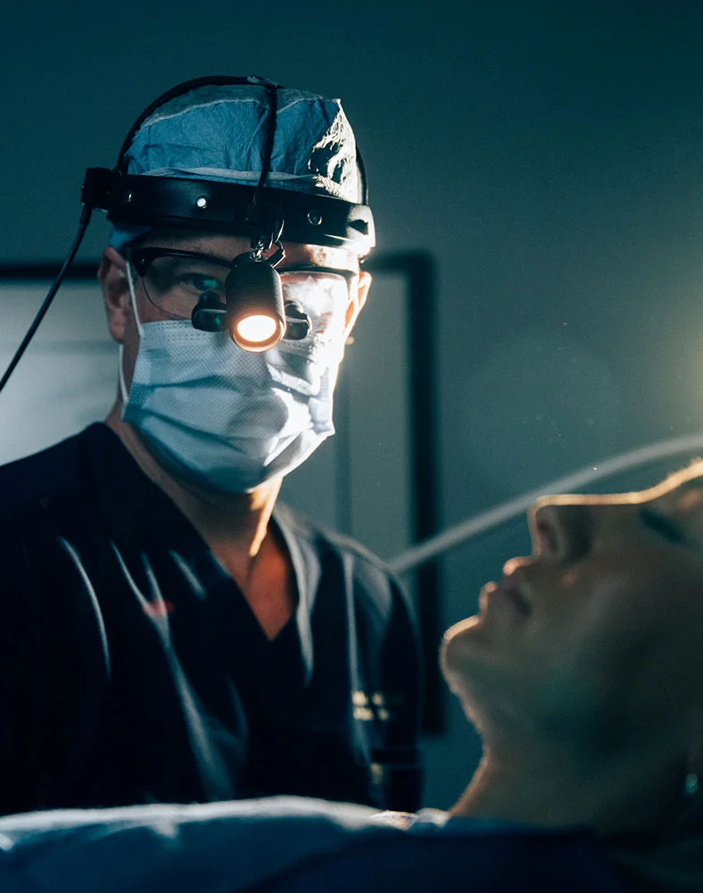 Close-up of a surgical scene featuring Dr. Hamilton. He is wearing a surgical cap, mask, and glasses equipped with a surgical light, intently focused on a patient lying on the surgical table. The patient’s profile and mouth area are partially visible. Focused lighting highlights the surgical site, creating deep shadows and emphasizing the clinical setting. The color palette includes muted tones of dark blue, gray, and soft light, conveying professionalism and precision. - Neck and Chin Liposuction in Carmel, IN