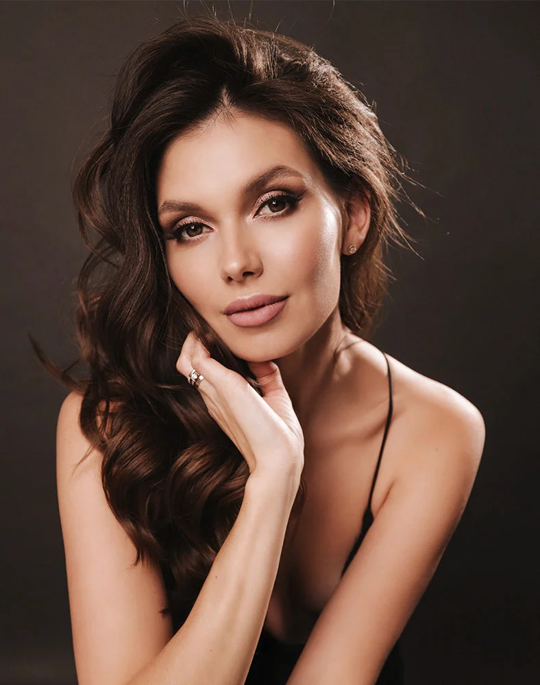 Close-up of a light-skinned woman with shoulder-length, wavy brown hair, seated with her head slightly tilted. She wears natural-toned makeup and a subtle blush, with warm brown eyes and a soft expression. Her hand, adorned with a delicate ring on the ring finger, rests gently on her cheek. She is dressed in a black sleeveless top or dress. The lighting is soft, and the dark background enhances the warmth of her features.- Neck Lift in Carmel, IN