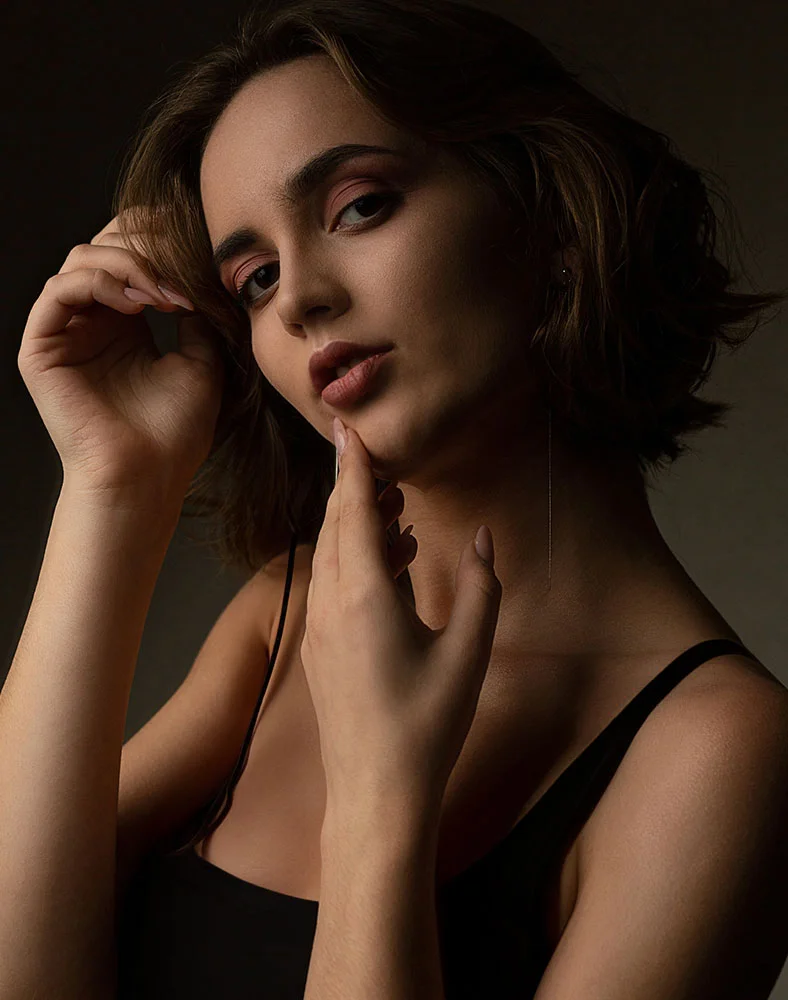 Close-up portrait of a young woman with shoulder-length, dark brown wavy hair. She wears subtle makeup with rosy tones and soft pink lips. Her expression is thoughtful, gaze slightly downward, with one hand resting gently on her face. She wears a black sleeveless top and thin gold earrings. Dramatic lighting highlights her features against a dark background. - Lip Augmentation in Carmel, IN