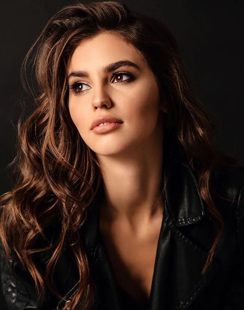 Close-up portrait of a woman with long, wavy dark brown hair, looking slightly upward and to her right with a thoughtful expression. She wears subtle makeup, including eyeshadow, eyeliner, mascara, and neutral lipstick. Her skin appears smooth and even-toned. She is dressed in a black leather jacket, and the dark background and soft, focused lighting highlight her face and upper body. - Sculptra in Carmel, IN
