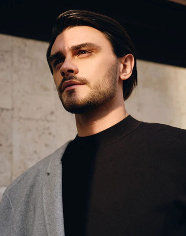 Close-up of a man with short dark brown hair and a well-groomed beard and mustache. He has a neutral expression and is looking slightly upward and off to the side. He is wearing a black turtleneck and a light gray wool jacket visible on the left side. The background features a light beige or taupe stone wall with natural lighting that casts soft shadows, suggesting an outdoor setting- Otoplasty in Carmel, IN