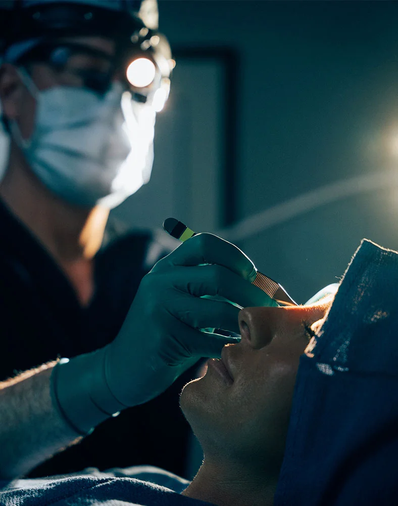 Surgeon Dr. Hamilton, wearing a surgical mask, glasses, and teal-colored gloves, performs a procedure focused on a patient’s eye. The surgeon’s hands hold a surgical instrument near the patient’s face. The patient lies in a surgical position, wearing a gown. Bright, focused lighting highlights the surgical area, while the background is blurred to emphasize the procedure. - Rhinoplasty in Carmel, IN
