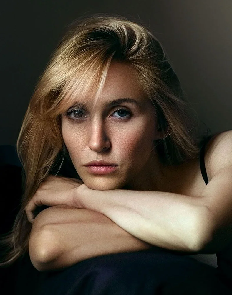 Close-up portrait of a woman with shoulder-length blonde hair styled in loose waves that fall over her face and shoulders. She has light-colored, expressive eyes and a neutral, slightly pensive expression, with her gaze directed slightly downward. Her arms are folded across her chest, resting on a dark fabric surface. Dramatic lighting highlights her features and casts soft shadows around her face and neck. The background is a deep, smoky-gray tone, creating a sophisticated and contemplative atmosphere. - Skin Cancer Reconstruction in Carmel, IN