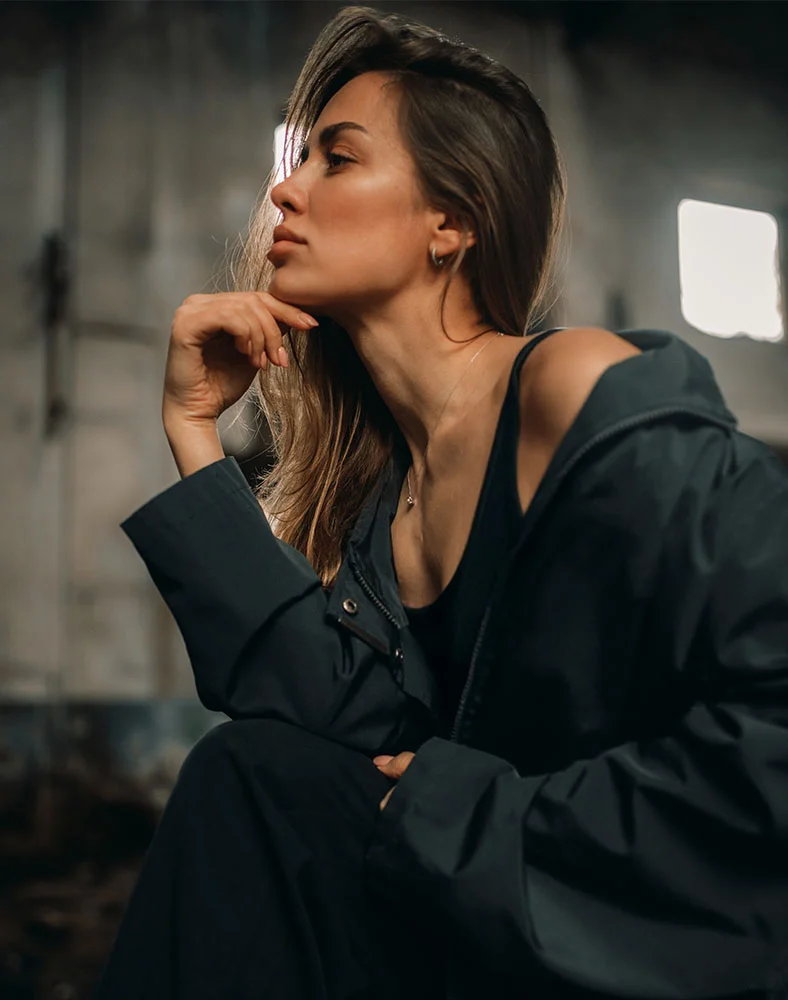 A woman with shoulder-length light brown hair is seated, angled slightly to her right, looking off to the right with her head slightly tilted. Her right hand rests on her chin in a thoughtful pose. She wears a dark gray or black long-sleeve windbreaker over a dark top. The muted gray background suggests an industrial or urban setting with soft shadows and diffused light, hinting at architectural details. - Submentoplasty in Carmel, IN